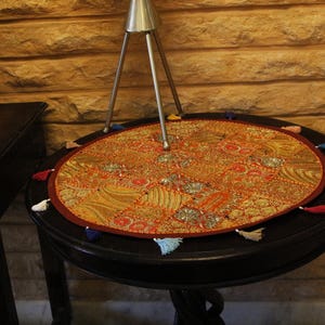 May include: An oval, decorative table mat with a detailed, colourful pattern in shades of orange, red, and gold. The mat is trimmed with a red border and small tassels in various colours. The mat rests on a dark, round table.
