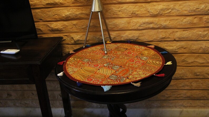 May include: An oval, decorative table mat with a detailed, colourful pattern in shades of orange, red, and gold. The mat is trimmed with a red border and small tassels in various colours. The mat rests on a dark, round table.