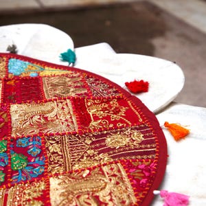 May include: A circular red textile floor cushion featuring a patchwork design of embroidered floral and geometric patterns in gold, green, and blue thread. The edge is decorated with small, colourful fabric tassels in teal, red, orange, and pink.