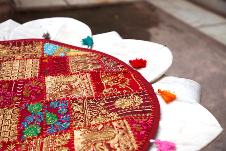May include: A circular red textile floor cushion featuring a patchwork design of embroidered floral and geometric patterns in gold, green, and blue thread. The edge is decorated with small, colourful fabric tassels in teal, red, orange, and pink.