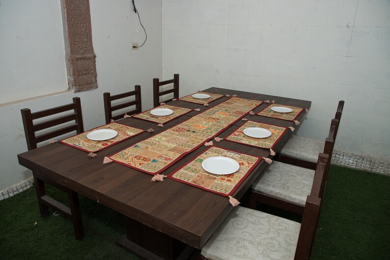 Puede incluir: Una mesa de comedor rectangular de madera marr&oacute;n oscuro con seis sillas a juego que tienen cojines de color claro. La mesa est&aacute; puesta con un camino de mesa rectangular de patchwork y seis manteles individuales a juego, cada uno con un plato blanco.