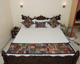 Puede incluir: Una cama con cabecero y piecero de madera tallada en color marrón oscuro, vestida con ropa de cama blanca. Incluye tres cojines cuadrados y un camino de cama rectangular largo, todos decorados con un diseño de patchwork multicolor.