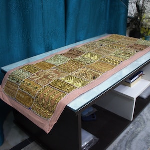 Könnte beinhalten: Rechteckiger Tischläufer mit Patchwork-Design. Der Läufer zeigt goldene, braune und grüne Stickmuster auf rosa Hintergrund. Der Läufer liegt auf einem dunkelbraunen Couchtisch. Der Tisch ist 120 cm lang.