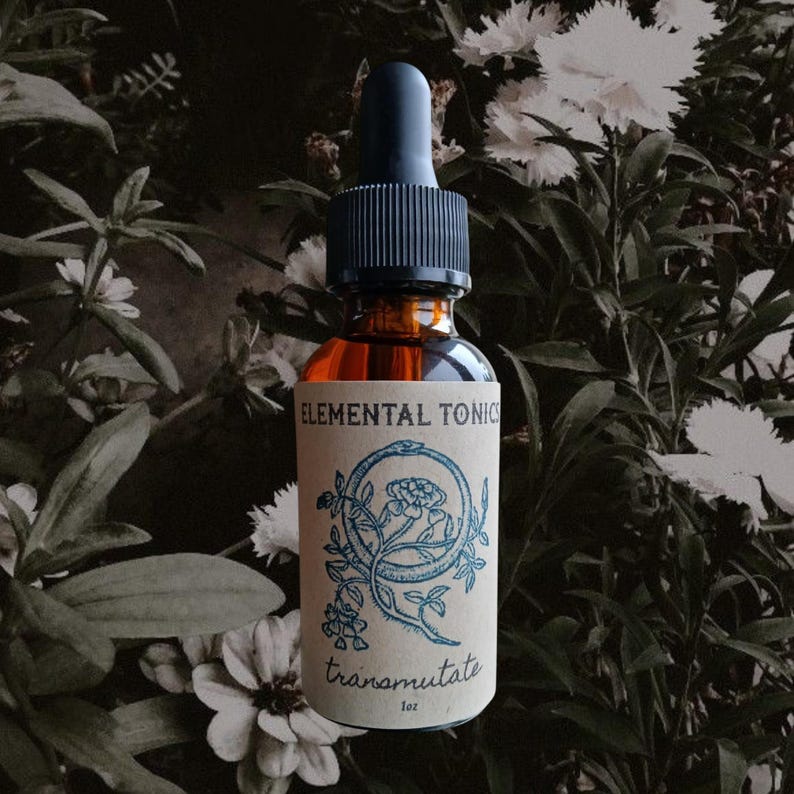 May include: A brown glass bottle with a black dropper top, labeled "ELEMENTAL TONICS" and "transmutate." The label features a blue illustration of a snake and flowers. The bottle contains a golden liquid and is set against a backdrop of green foliage and white flowers.