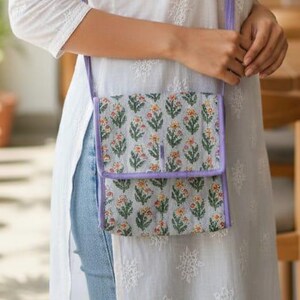 以下が含まれることがあります： 白い刺繍チュニックと水色のジーンズ。緑、オレンジ、黄色の花柄の小さな長方形のクロスボディバッグが肩からかけてあります。バッグはラベンダー色のストラップとトリムが付いています。