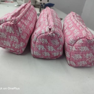 Może przedstawiać: Trzy różowe, pikowane kosmetyczki z białym wzorem słoni. Każda torba ma zapięcie na zamek błyskawiczny i zaokrąglony kształt. Torby są wykonane z miękkiej tkaniny i prawdopodobnie ręcznie robione.