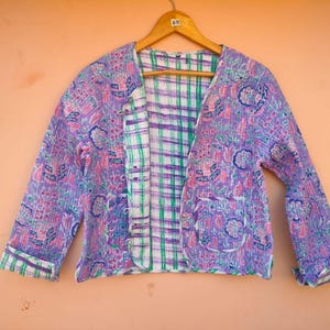 May include: A reversible jacket with a floral pattern in shades of purple, pink, and blue. The jacket has a plaid lining in green, white, and purple. It is hanging on a wooden hanger against a peach-colored background.