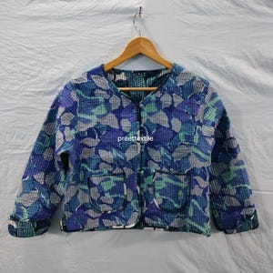 May include: A blue, green, and gray patterned jacket with a round collar and two front pockets. The jacket is hanging on a wooden hanger against a white background. The jacket has a quilted texture.