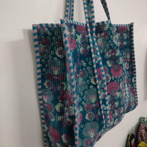 Puede incluir: Un bolso tote grande con un estampado floral en tonos turquesa, rosa y verde. El bolso tiene asas y ribetes turquesa con un dise&ntilde;o a rayas. El bolso est&aacute; hecho de una tela texturizada y est&aacute; colgado contra una pared blanca.