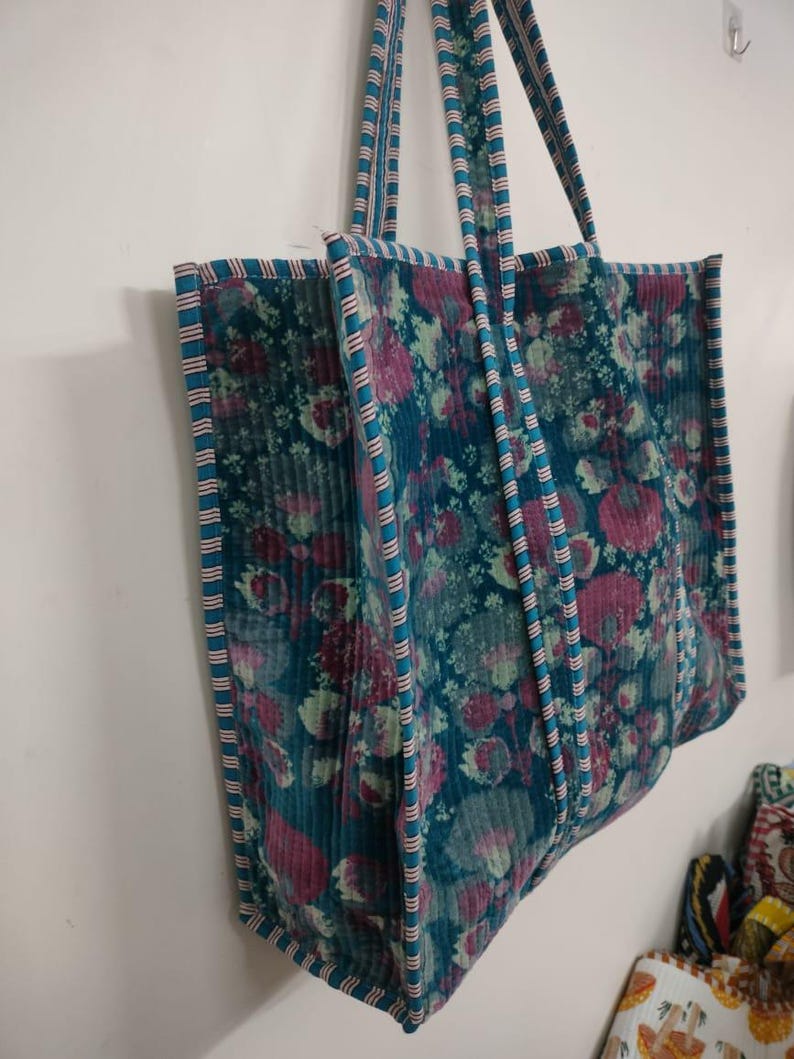 Puede incluir: Un bolso tote grande con un estampado floral en tonos turquesa, rosa y verde. El bolso tiene asas y ribetes turquesa con un dise&ntilde;o a rayas. El bolso est&aacute; hecho de una tela texturizada y est&aacute; colgado contra una pared blanca.