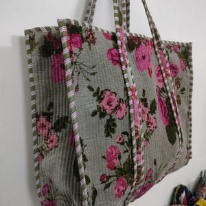 Puede incluir: Un bolso tote grande con un estampado floral. El bolso presenta un fondo gris con rosas rosas y hojas verdes. Los bordes y las correas est&aacute;n ribeteados con tela a rayas verdes y rosas. El bolso cuelga contra una pared blanca.