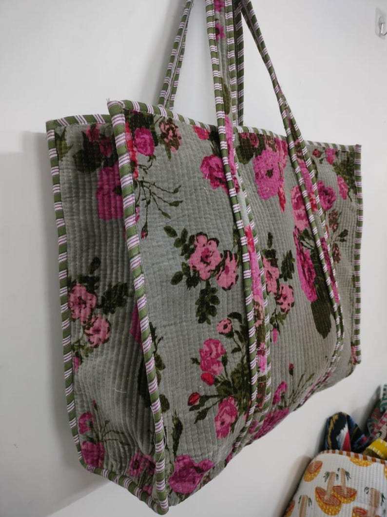 Puede incluir: Un bolso tote grande con un estampado floral. El bolso presenta un fondo gris con rosas rosas y hojas verdes. Los bordes y las correas est&aacute;n ribeteados con tela a rayas verdes y rosas. El bolso cuelga contra una pared blanca.