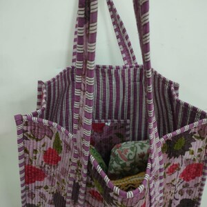 Puede incluir: Un bolso tote acolchado con un estampado floral en tonos morados, rojos y verdes. El bolso tiene un forro y asas a rayas. El bolso tiene m&uacute;ltiples bolsillos y est&aacute; dise&ntilde;ado para transportar objetos.