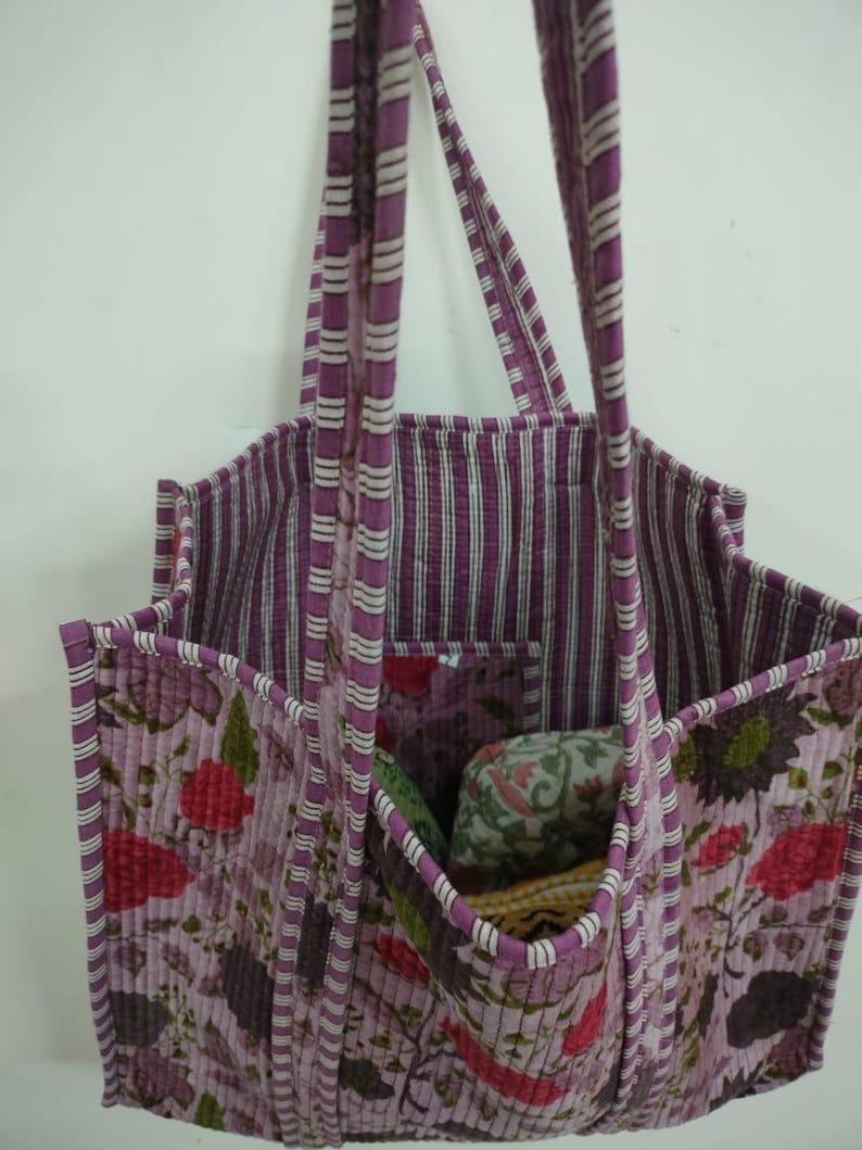 Puede incluir: Un bolso tote acolchado con un estampado floral en tonos morados, rojos y verdes. El bolso tiene un forro y asas a rayas. El bolso tiene m&uacute;ltiples bolsillos y est&aacute; dise&ntilde;ado para transportar objetos.
