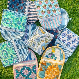 Puede incluir: Una colección de coloridas bolsas acolchadas con varios patrones florales y geométricos. Cada bolsa tiene un bolsillo con cremallera y está hecha de tela estampada en tonos de azul, rosa, verde y amarillo. Las bolsas están dispuestas sobre hierba verde.