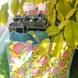 May include: A collection of tote bags with various floral and striped patterns. The bags feature a yellow floral print with pink flowers and green leaves, a blue and white striped pattern, and a black and white striped pattern. The bags have handles and are displayed on a grassy surface.