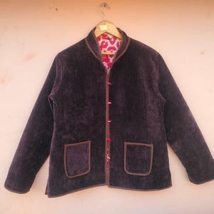 Op de afbeelding: Een donkerbruine corduroy jas met een opstaande kraag, twee voorzakken en een patroon aan de binnenkant. De jas heeft een knoopsluiting en bruine biezen. De jas hangt aan een houten kleerhanger.