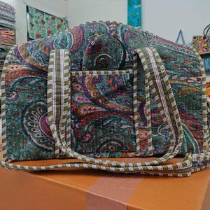 Puede incluir: Un bolso tote acolchado con estampado de cachemira en tonos verdes, morados y rosas. El bolso tiene una pequeña bolsa a juego y correas largas. Las correas y el ribete tienen un patrón de rayas blancas y verde oliva. El bolso está sobre una superficie marrón.