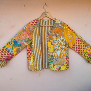 Op de afbeelding: Een patchworkjas met diverse bloemen- en geometrische patronen in geel, oranje en roze tinten. De jas heeft lange mouwen en een gestreepte binnenkant. De jas hangt aan een houten hanger.