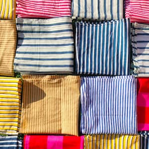 Pode incluir: Vista aérea de quadrados de tecido dobrados em várias cores e padrões listrados. As cores incluem amarelo, bege, rosa, vermelho, azul e branco. Os tecidos são dispostos em um padrão de grade, criando uma exibição vibrante e texturizada.