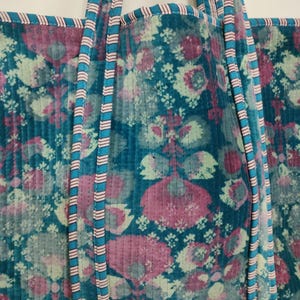Puede incluir: Un bolso tote acolchado con un estampado floral en tonos turquesa, rosa y blanco. El bolso tiene correas turquesas con rayas rosas y blancas. Los bordes del bolso est&aacute;n ribeteados con tela rosa.