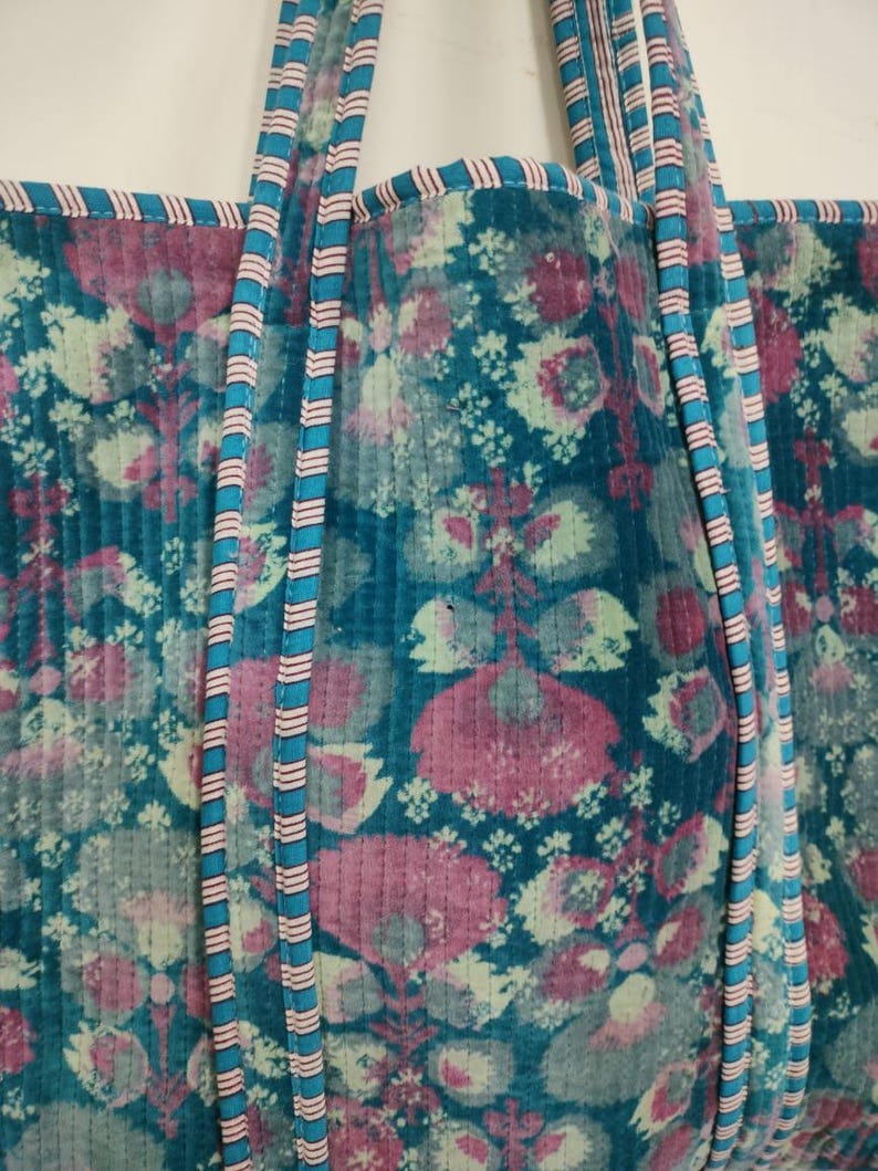 Puede incluir: Un bolso tote acolchado con un estampado floral en tonos turquesa, rosa y blanco. El bolso tiene correas turquesas con rayas rosas y blancas. Los bordes del bolso est&aacute;n ribeteados con tela rosa.