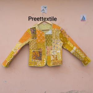 May include: A patchwork jacket in shades of yellow, orange, and green, with various patterned fabric squares. The jacket is hanging on a wooden hanger against a light pink background. The text "Preettextile" is visible.