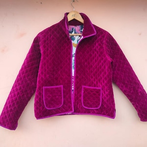 May include: A quilted magenta jacket with a collar and two front pockets. The jacket has a diamond pattern and is lined with a floral print. The edges of the jacket and pockets are trimmed with a darker magenta color.