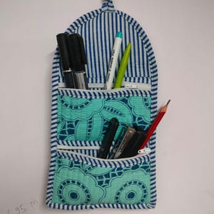 May include: A quilted fabric wall organizer with three pockets. The organizer features a blue and white striped top section and two turquoise pockets with a floral pattern. Various pens and pencils are stored inside.