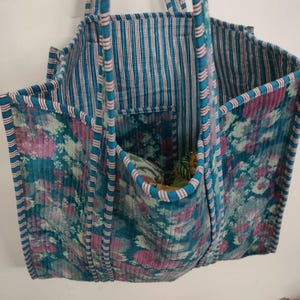 Puede incluir: Un bolso tote acolchado con un estampado floral en verde azulado, rosa y blanco. El bolso tiene un interior y asas a rayas, con un bolsillo en la parte delantera. El bolso est&aacute; hecho de una tela acolchada suave.