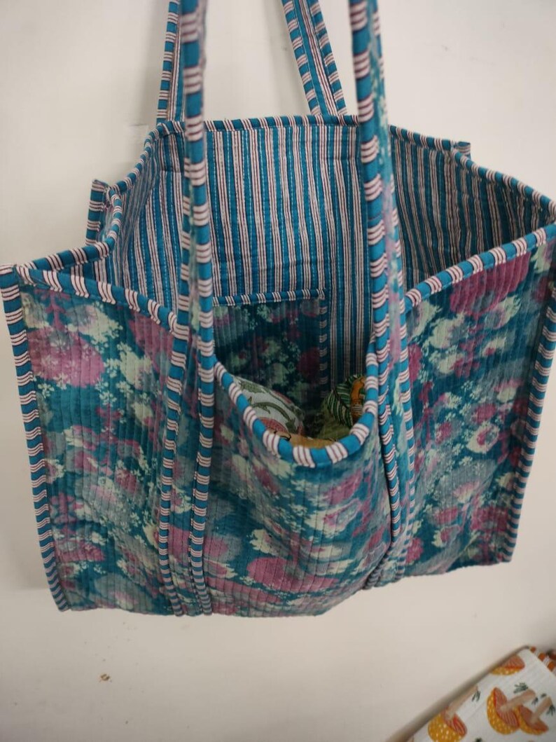 Puede incluir: Un bolso tote acolchado con un estampado floral en verde azulado, rosa y blanco. El bolso tiene un interior y asas a rayas, con un bolsillo en la parte delantera. El bolso est&aacute; hecho de una tela acolchada suave.