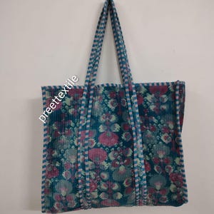 Puede incluir: Un bolso tote grande con un estampado floral en tonos turquesa, rosa y blanco. El bolso tiene asas largas y el texto "preette textile" es visible en el lateral. El bolso est&aacute; colgado contra una pared blanca.
