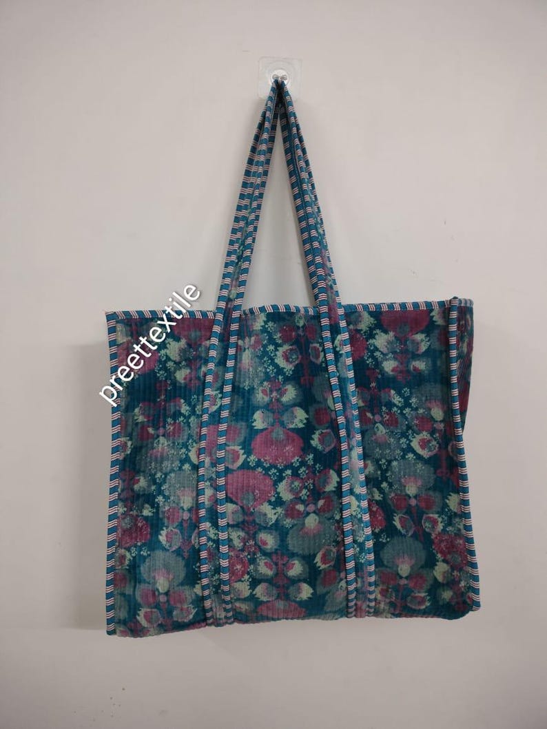 Puede incluir: Un bolso tote grande con un estampado floral en tonos turquesa, rosa y blanco. El bolso tiene asas largas y el texto "preette textile" es visible en el lateral. El bolso est&aacute; colgado contra una pared blanca.