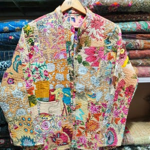 May include: A quilted jacket with a vibrant floral patchwork design. The jacket features a mandarin collar, wooden buttons, and a front pocket. The color palette includes shades of pink, orange, yellow, and green, creating a bohemian aesthetic.