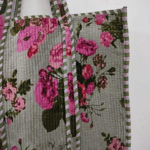 Puede incluir: Un bolso tote acolchado con un estampado floral de rosas rosas sobre un fondo verde apagado. El bolso tiene ribetes a rayas verdes y rosas y asas a juego. La tela tiene una apariencia acolchada y texturizada.