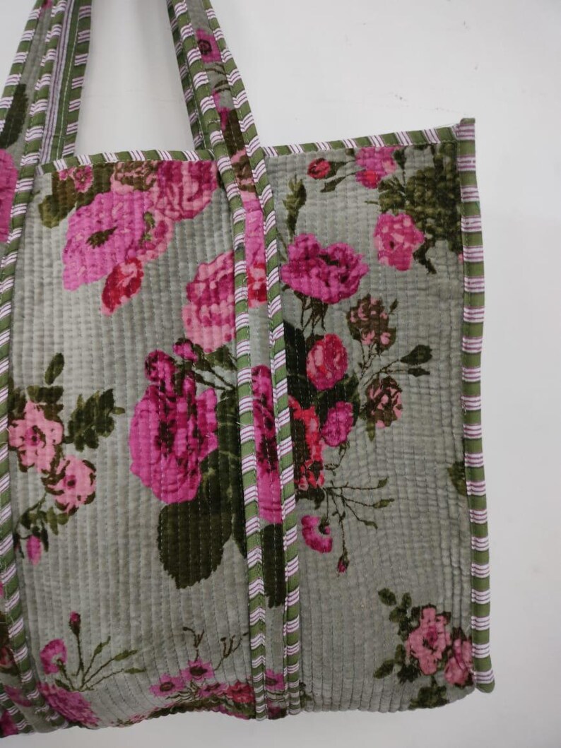 Puede incluir: Un bolso tote acolchado con un estampado floral de rosas rosas sobre un fondo verde apagado. El bolso tiene ribetes a rayas verdes y rosas y asas a juego. La tela tiene una apariencia acolchada y texturizada.
