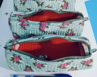 Puede incluir: Tres estuches florales apilados, cada uno con un exterior acolchado azul claro adornado con flores rosas. Los interiores son de un rojo vibrante. Un bolígrafo azul descansa delante, lo que sugiere su uso para artículos de papelería o pequeños objetos.