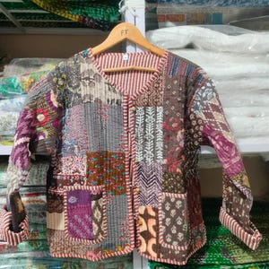 Puede incluir: Chaqueta patchwork con una variedad de cuadrados de tela estampada en tonos marrones, grises, morados y rojos. La chaqueta tiene mangas largas, cuello en V y dos bolsillos delanteros. La chaqueta está colgada en una percha de madera.