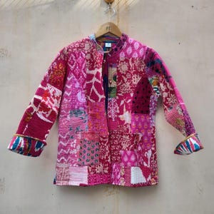 Peut inclure: Veste patchwork aux tons de rose, magenta et blanc, suspendue à un cintre en bois. La veste présente une variété de motifs floraux et géométriques, avec des poignets contrastés. C'est un vêtement unique.
