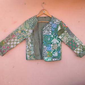 May include: A patchwork jacket with a variety of green and white floral and geometric patterns. The jacket has long sleeves and a striped interior. It is hanging on a wooden hanger against a pink background.