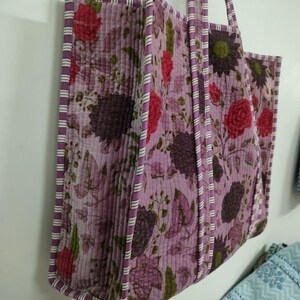Puede incluir: Un bolso tote acolchado con un estampado floral en tonos morados, rojos y verdes. El bolso tiene forma rectangular con un borde y asas a rayas. El bolso est&aacute; colgado en una pared blanca.
