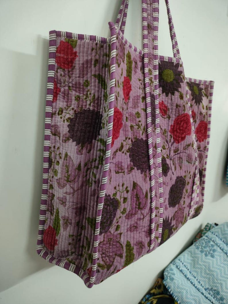 Puede incluir: Un bolso tote acolchado con un estampado floral en tonos morados, rojos y verdes. El bolso tiene forma rectangular con un borde y asas a rayas. El bolso est&aacute; colgado en una pared blanca.