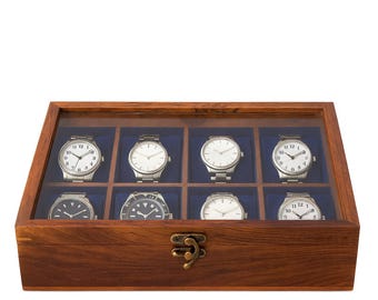 May include: A wooden watch display case with a clear glass top, showcasing eight wristwatches. The watches have silver-toned cases and various white or black watch faces. The case is made of dark wood and has a brass-colored clasp.