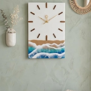 May include: A rectangular wall clock with a beach-themed design. The clock face is white with brown wooden markers and hands. The bottom half features a depiction of a beach with blue and white waves. The clock measures approximately 12 inches by 18 inches.