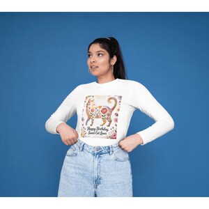 Puede incluir: Camiseta blanca de manga larga con un diseño floral de gato. El gato está decorado con flores y hojas rosas, amarillas y verdes. El texto "Happy Birthday Sweet Cat Lover" está debajo.