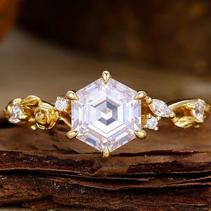 May include: A gold ring with a large, hexagonal, clear gemstone as the centerpiece. The ring features smaller, clear accent stones and floral detailing on the band. The ring is set on a piece of dark brown wood.