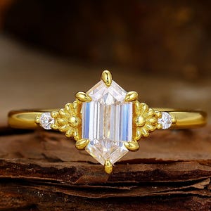 May include: A gold ring featuring a large, rectangular, clear gemstone with a faceted cut. The ring is accented with floral designs and small, round gemstones on either side of the main stone. The ring is set on a wooden surface.