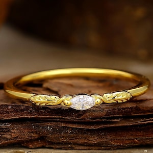 Könnte beinhalten: Ein goldener Ring mit einem Diamanten im Marquiseschliff. Das dünne Band hat blattartige Muster auf beiden Seiten des Diamanten. Der Ring liegt auf dunklem, braunem Holz.