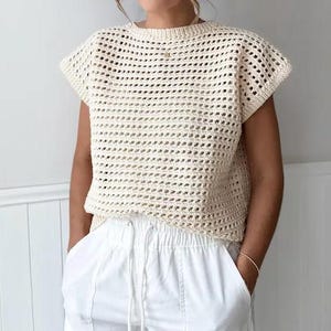 Könnte beinhalten: Ein cremefarbenes Kurzarm-Oberteil mit einem offenen Häkelmuster und einem Rundhalsausschnitt. Das Kleidungsstück wird mit einer weißen Hose kombiniert, die einen Kordelzug in der Taille und sichtbare vertikale Nähte auf der Vorderseite hat.