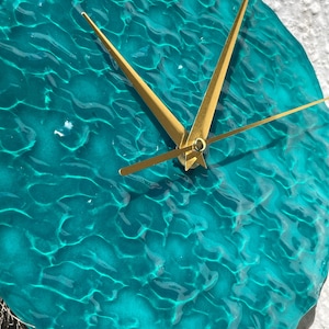 May include: A circular wall clock with a translucent teal face featuring a textured, rippled surface that mimics the appearance of water. The clock has three thin, gold-colored metal hands centered on the face, indicating the time.